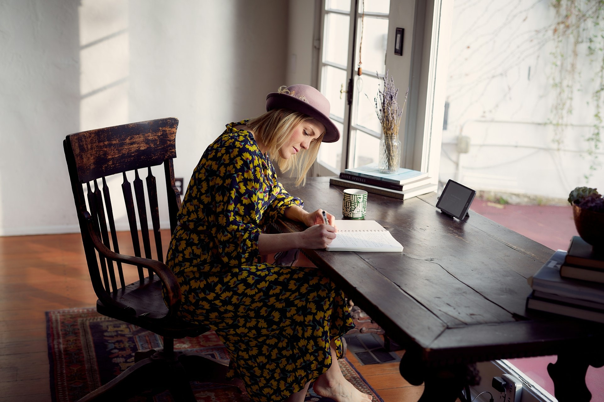 A stock image of a woman wearing a dress and hat, sitting at a desk writing in a notebook. This is to illustrate the topic of getting paid to write poetry