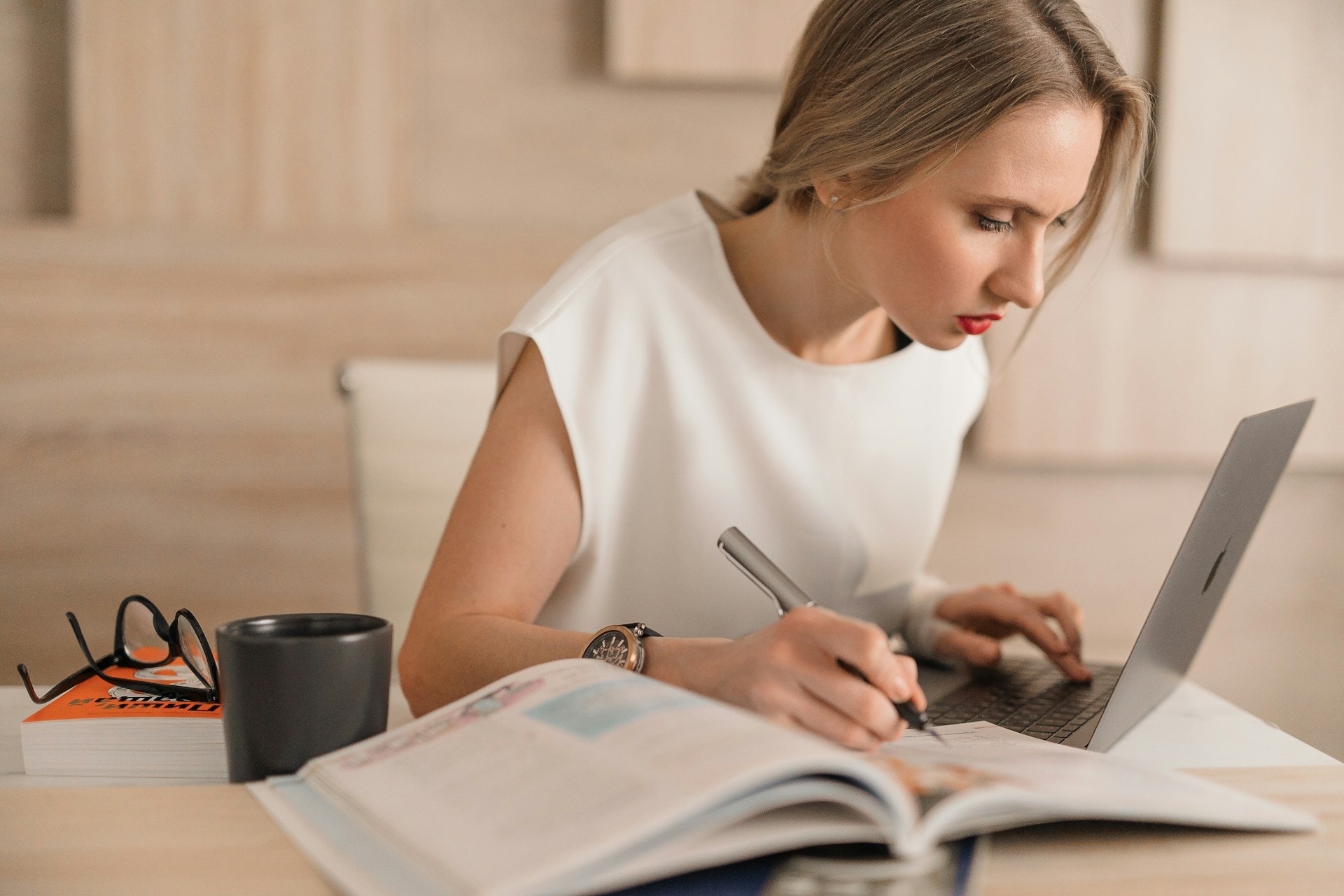 An AI generated image of a woman looking at a laptop while holding a pen over a textbook. This is to emphasize that finding freelance Christian writing jobs has never been easier
