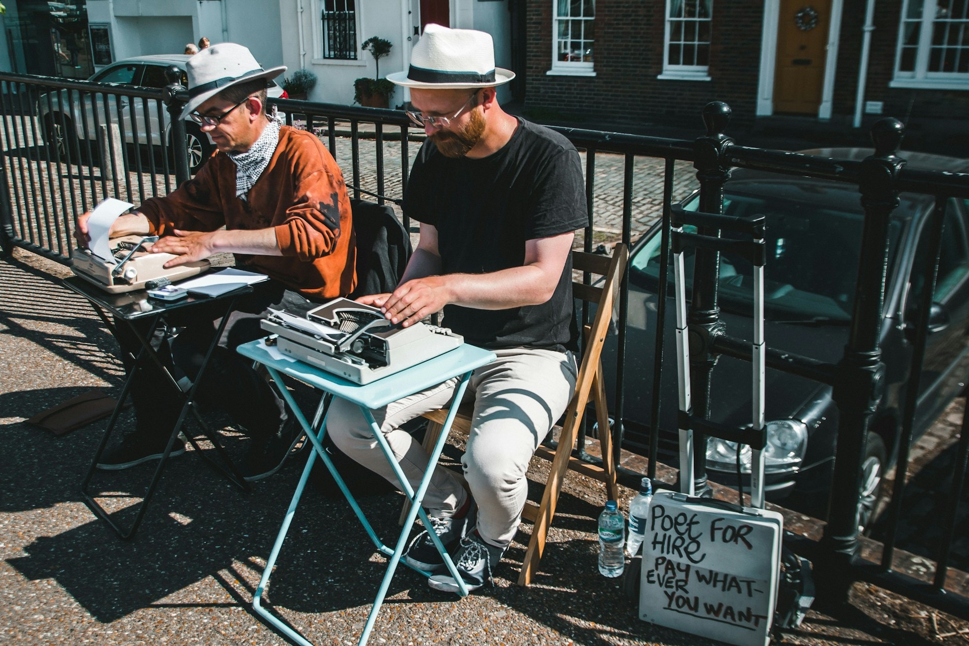 A stock image of two men sitting outside on chairs with small TV tables holding a typewriter in front of them. They are wearing stereotypically beatnik clothing and hats. A nearby suitcase reads "Poet for hire. Pay whatever you want." This is to illustrate how people can make money writing poetry today!